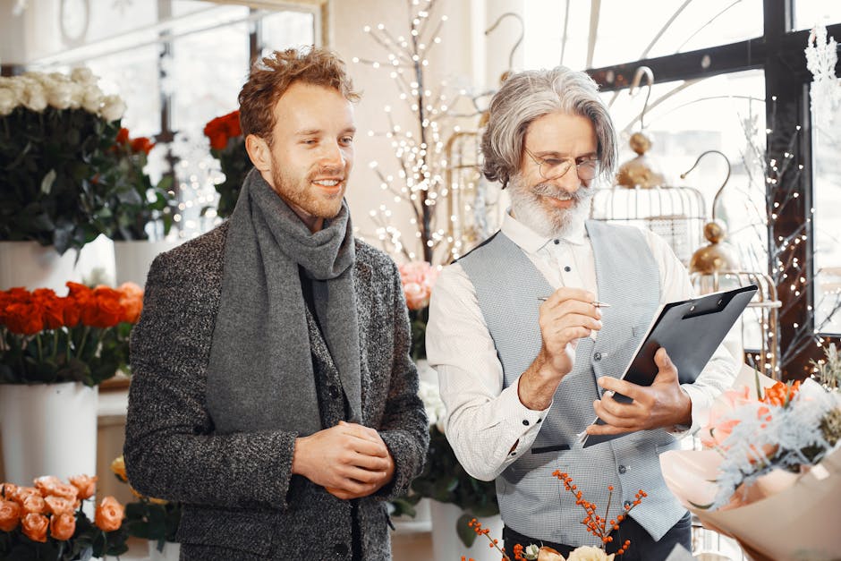 A scene inside a bright flower shop showing a young man with light brown hair, wearing a dark textured coat and a grey scarf, standing next to an older man with grey hair, glasses, and a beard, dresse