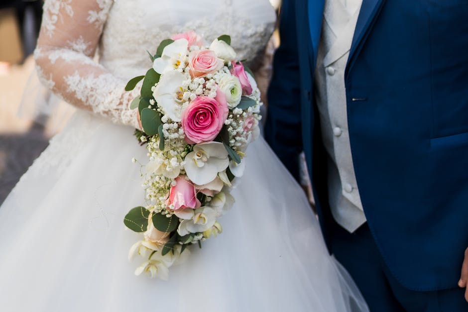 Close-up of a bridal bouquet featuring pink roses, white orchids, and baby's breath, accented with green eucalyptus leaves, held by a bride dressed in a lace wedding gown with long sleeves. The bouque