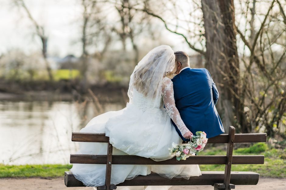 A bride and groom sit together on a wooden park bench near a calm river, with the bride wearing a white wedding gown with lace sleeves and a flowing veil, and the groom in a navy blue suit. The bride 